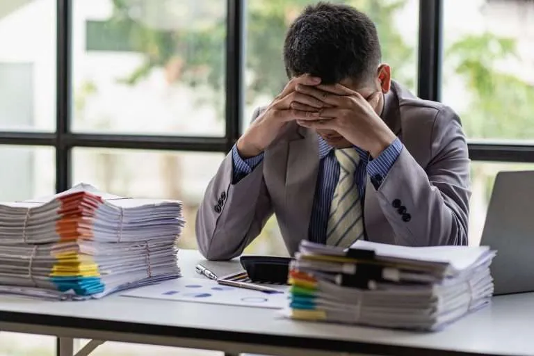 A busy businessman sleeps at his desk with his financial calculators and papers piled up on a pile of desks with laptops.