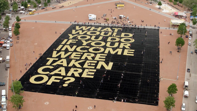 Aerial view of a public square with a large sign on the ground saying "What would you do if your income were taken care of?"