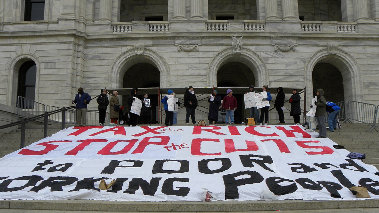 Protestors in front of a building with a large sign advocating to tax the rich.